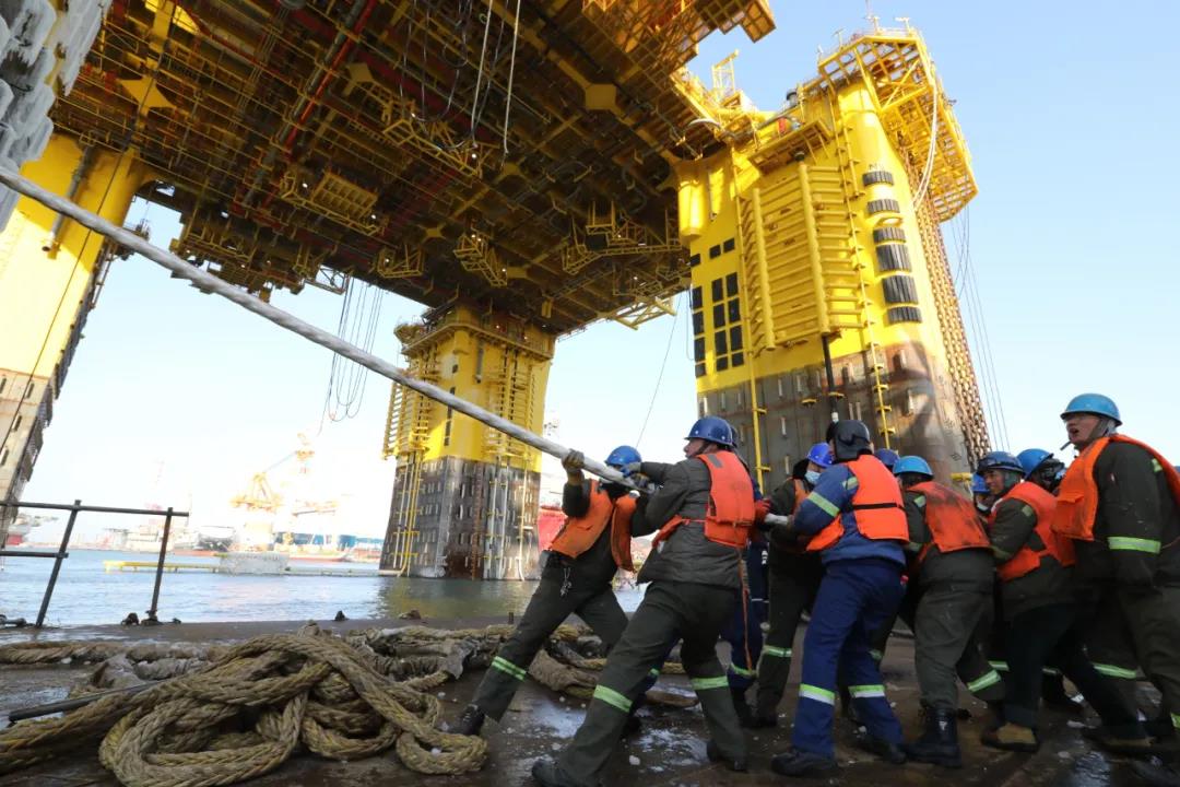 紅日剛剛探出頭，施工人員已經聚集在碼頭，準備解纜。每四五個工人駕駛一條小漁船，在船體立柱下拉開粗粗的纜繩，岸上的工人則排成一列，像拔河一樣有節奏地奮力向后拉。拖輪靠近船體立柱，工人們奮力拉拽沉重的纜繩，進行帶纜作業。