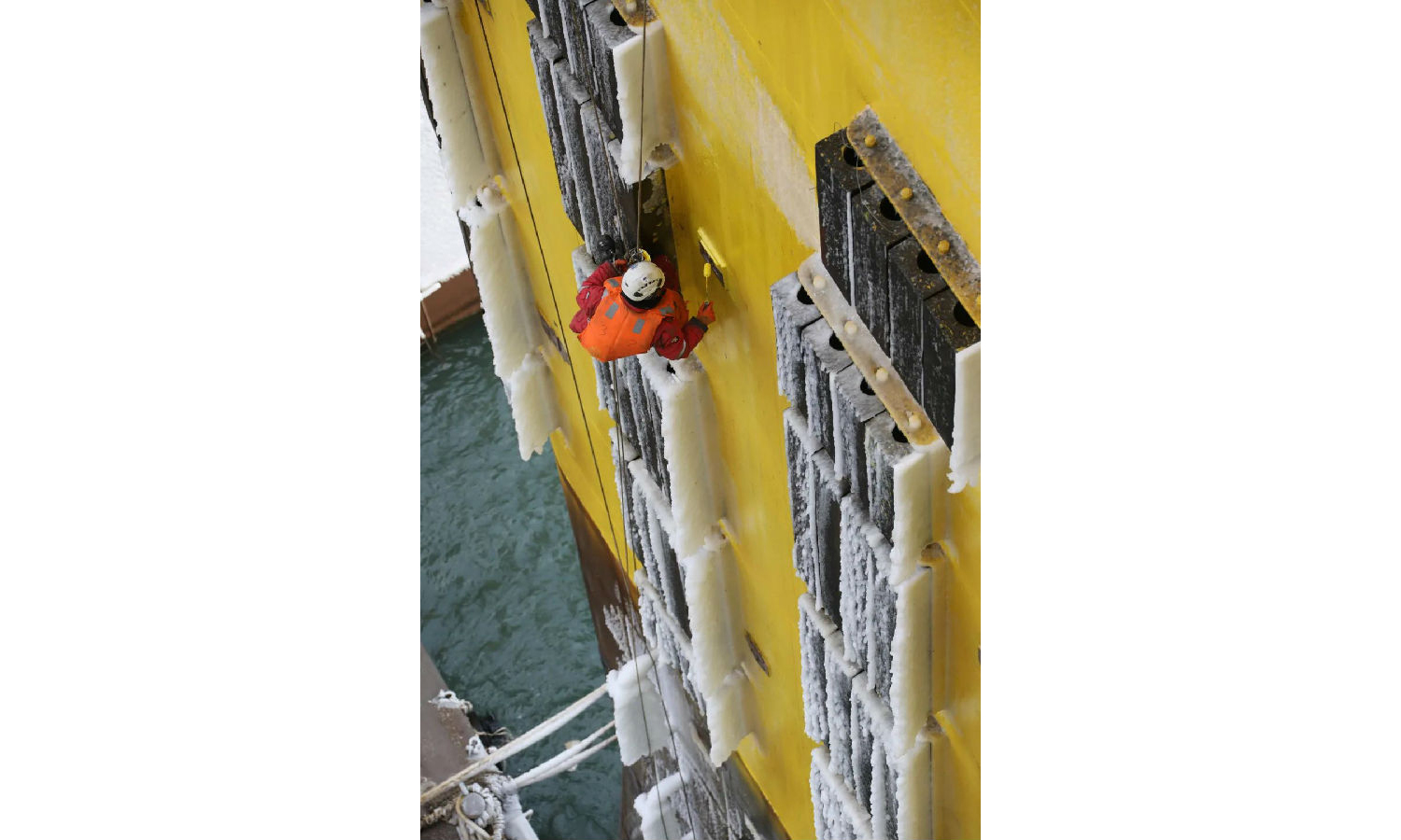 在芝罘灣冬日的風雪中，工作人員裹緊棉工裝，吊籠在空中緩慢旋轉升降，平臺上的水印凝成厚厚的冰霜，飯菜吃一半就涼，生活樓取暖全靠“小太陽”。圖為“蜘蛛人”懸掛在高空進行船體立柱補漆作業，為啟航做好準備。