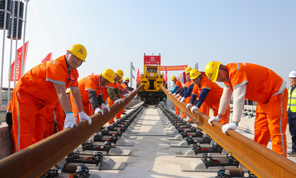 為優質高效完成鋪軌任務，建設者提前在位于荊州站北側的鋪軌基地儲存長鋼軌，以鋪軌基地為起點，采用TC-500型鋪軌機由荊州站一路向北鋪設長鋼軌；加持智能行車調度指揮系統，全天候對鋪軌機、長軌運輸車、焊軌車等設備的運行狀態及作業位置進行監控和統一調度指揮，保證了交叉施工安全有序，并且提高了鋪軌速度，每天單向鋪軌可達6公里。圖為鋪軌作業現場，作業人員在鋪軌機的牽引下正在安裝軌道。