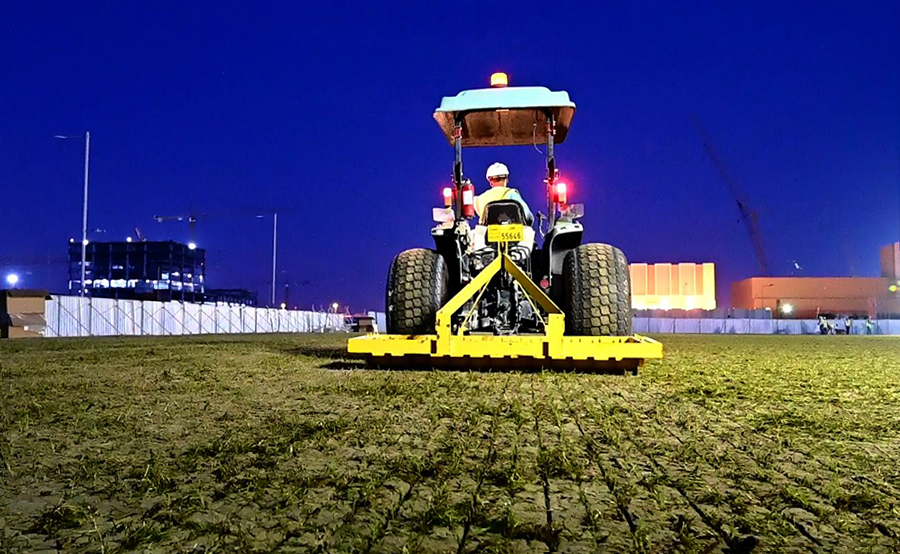 項目部細心策劃場地平整、播種、灌溉等環節，以保障建植質量。為避免白天高溫影響草皮種植，建設者們在夜間完成了播種、灌溉及覆砂等工作。圖為工作人員操作扎草機把小草根莖扎進土地。